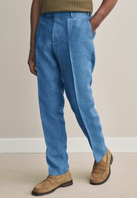 Lichtblauwe linnen broek met een getailleerde pasvorm, voorzien van twee zijzakken en een vouw aan de voorkant. Gecombineerd met bruine suède schoenen.