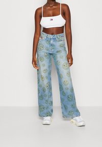 Žena v světle modrých širokých džínech s potiskem zelených usmívajících se obličejů, bílé crop top a bílé tenisky, stojící na jednobarevném pozadí.