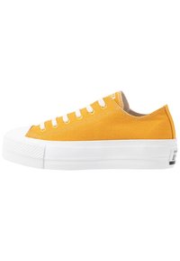 Keltainen kangaskenkä valkoisella kumipohjalla, pyöreä kärki ja kuusi valkoista nauharengasta. Ominaisuuksiin kuuluu teksturoitu kärkipaneeli ja merkkilogo kantapäässä.