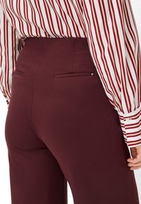 Pantaloni bordeaux realizzati in tessuto liscio, con una silhouette aderente e due tasche posteriori, abbinati a una blusa a righe verticali.