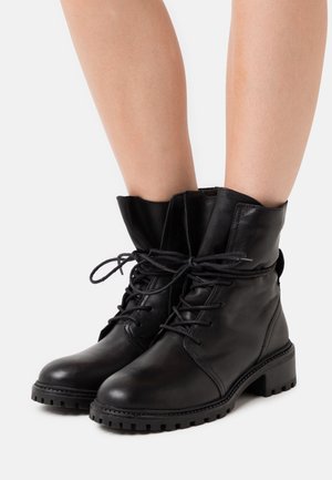 Bottes basses en cuir noir avec un bout arrondi, un laçage à l'avant et une semelle en caoutchouc texturée. Détail de col légèrement replié à l'ouverture.