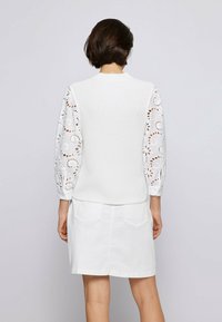 Kvinna med kort brunt hår iförd en vit stickad väst över en långärmad broderad blus och en vit kjol, vänd bort från kameran.