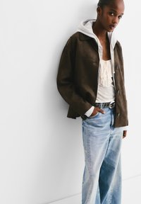 Veste en daim marron avec une capuche grise, portée sur un haut blanc froncé. Associée à un jean à jambes larges bleu clair et une ceinture décorative.