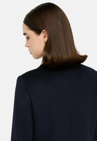 Blazer blu navy con risvolti a lancia, tessuto liscio e vestibilità su misura, indossato da una persona vista di lato e di schiena, con capelli lisci e dritti.