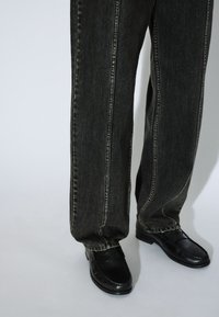 Zwarte spijkerbroek met een losse pasvorm, met contrasterende stiksels langs de zijkanten, gecombineerd met zwarte lederen schoenen.