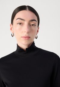 Zwarte hoogsluitende top met een soepele textuur. Model draagt zilveren oorringen. Eenvoudig, minimalistisch ontwerp.