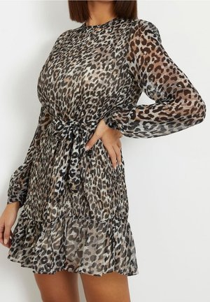 Femme portant une robe longue à manches longues, en tissu transparent à imprimé léopard, avec une ceinture à la taille et un ourlet à volants, posant avec la main sur la hanche devant un fond uni.