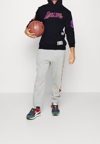 Felpa nera con la scritta "LAKERS", pantaloni della tuta grigi con grafiche laterali e scarpe da ginnastica multicolori. Tiene un pallone da basket.