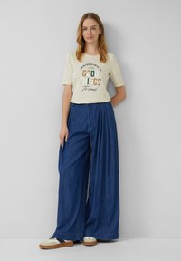 T-shirt crème à manches courtes avec texte imprimé ; large pantalon en denim bleu ; baskets beiges et noires. Tissu lisse avec une coupe relaxée.