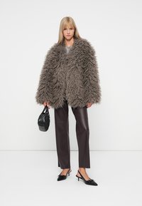 Καφέ χνουδωτό ψεύτικο μπουφάν με φαρδιά μανίκια, συνδυασμένο με σκοτεινές καφέ ψεύτικες δερμάτινες παντελόνες και μαύρες γόβες με μυτερή μύτη, κρατώντας μια μικρή τσάντα.