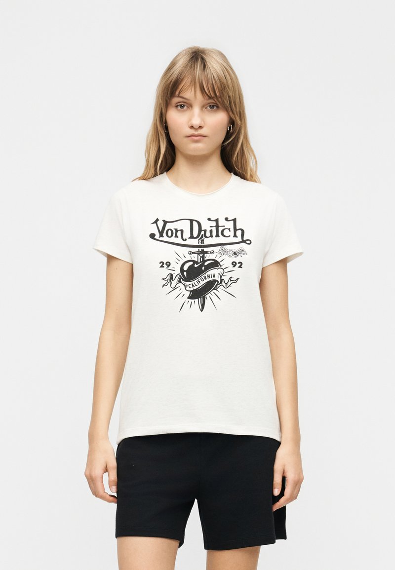 Νεαρή γυναίκα που φοράει λευκό t-shirt Von Dutch με μαύρο γραφικό και κείμενο, συνδυασμένο με μαύρο σορτς, στέκεται μπροστά σε απλό φόντο.