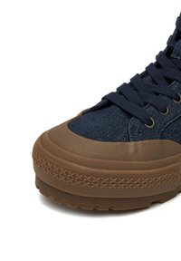 Tumši zilas kanvas augstpapēžu kurpes ar brūnu gumijas zolīte. Tām ir fakturēts augšdaļa, apaļa purngala forma un metāla acuļi šņoreēm.