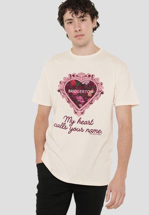 Young man wearing cream t-shirt with a floral heart design labeled "Bridgerton" and text "My heart calls your name."