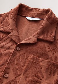 Chemise texturée marron avec col, ornée de motifs en relief représentant une main et un soleil, deux boutons et une petite poche poitrine. Détail du tissu doux.
