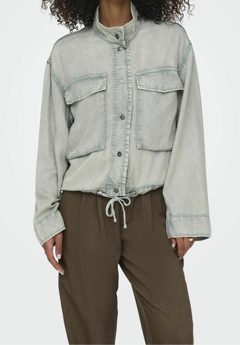 Femme portant une veste gris clair boutonnée avec deux poches poitrine et un pantalon marron ample, debout les mains détendues.