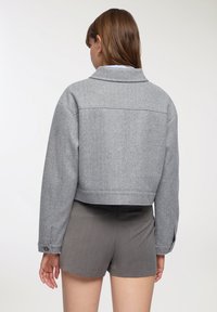 Kobieta z brązowymi włosami nosi jasnoszary krótki płaszcz i ciemnoszare szorty, stojąc tyłem do kamery na jednolitym jasnym tle.