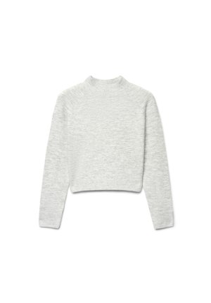 Giovane donna in piedi che sorride, indossa un maglione grigio chiaro, pantaloni ampi grigio scuro e scarpe da ginnastica bianche su sfondo bianco.