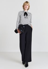 Schwarze Hosen mit weitem Bein, kombiniert mit einer weißen Bluse mit Muster und einem schwarzen Bindeband am Hals. Das Modell hält eine schwarze Clutch in der Hand.