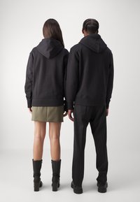 Dve osobe stoje leđima okrenute jedna drugoj, obučene u usklađene crne dukserice s kapuljačom. Dukserice su jednobojne, s rebrastim manžetama i opuštenim krojem.