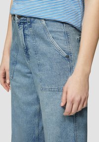 Pantalon en denim bleu clair avec une coupe décontractée. Comprend des poches avant, des coutures visibles et une fermeture à bouton. Texture lisse.