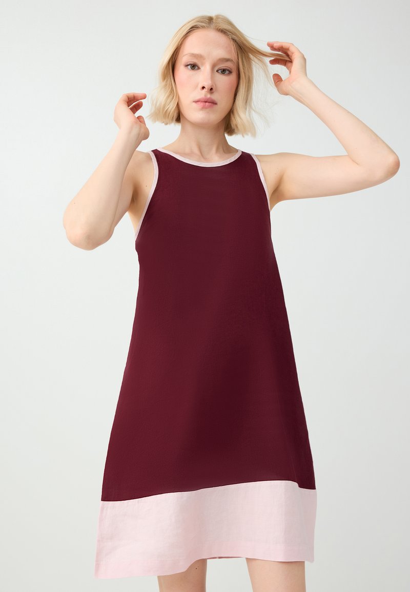 Femme blonde portant une robe bordeaux sans manches avec un ourlet rose clair, levant les mains près de sa tête sur un fond clair uni.