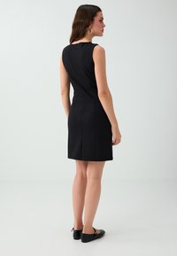 Woman with long brown hair wearing a sleeveless black knee-length dress and black flats, standing and facing slightly right.