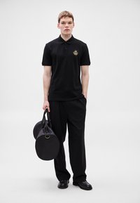 Melna polo krekls ar zeltainiem izšuvumiem, kopā ar vaļīgām melnām biksēm. Modelim rokās divas apaļas melnas somas ar rokturiem.