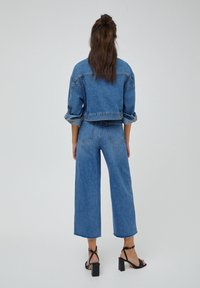 Denkeljacke und weite Jeans im mittleren Blau. Die Jacke hat aufgerollte Ärmel, während die Jeans einen rohen Saum und Gesäßtaschen besitzen.