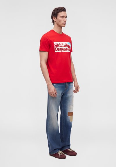 Мъжки модел стои пред обикновен фон, облечен в червена тениска Ski Club Dsquared2, широки сини дънки и бордо маратонки.