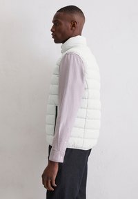 Gepolsterte weiße Weste mit hohem Kragen und seitlichen Reißverschlusstaschen, getragen über einem hellviolett gestreiften Hemd, seitlich gezeigt vor einem neutralen Hintergrund.