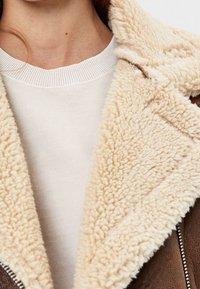Collo in shearling marrone con fodera in finta pelliccia crema, indossato sopra un maglione beige chiaro con scollo a costine.