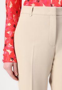 Pantalons taillés beiges avec une texture lisse, dotés d'un devant plat et de poches latérales. Associés à un haut à motifs floraux vibrants.