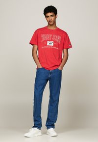 Camiseta de algodón roja con el texto "TOMMY JEANS", de manga corta, combinada con jeans de mezclilla azul y zapatillas blancas. Ajuste simple y casual.