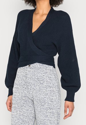 Femme portant un pull cache-cœur en maille côtelée bleu marine avec des manches ballon et un pantalon blanc à motifs taille haute.
