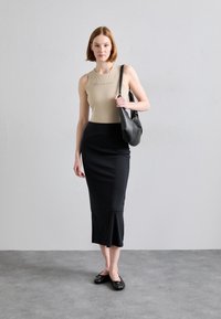 Beige mouwloos topje, zwarte aansluitende midi rok, zwarte platte schoenen. Model houdt een zwarte schoudertas vast en staat tegen een neutrale achtergrond.
