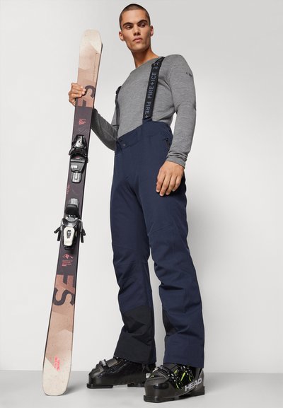 Marineblaue Skihosen mit verstellbaren Trägern und Taschen, graues Langarmshirt, hält ein Paar Skier mit schwarzen Bindungen und einer gemusterten Spitze.