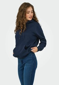 Hoodie bleu marine en matériau doux, avec une poche avant, une capuche à cordon de serrage et des poignets côtelés. Associé à un jean bleu ajusté.