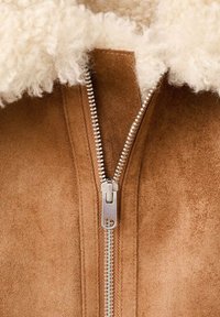 Bruine suède jas met een zilveren ritssluiting en een witte schapenvachtkraag. De jas heeft een zachte textuur en een close-up van de ritssluiting.