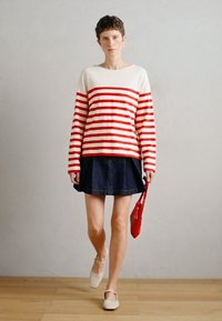 Sarkans un balts svītrots garo piedurkņu krekls, kas saskaņots ar tumši zilu denima svārku. Modelis tur nelielu sarkanu somiņu un valkā bēšas kurpes.