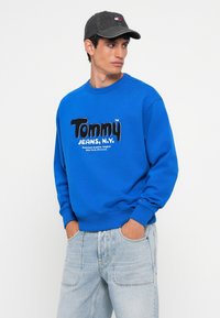 Felpa in cotone blu con la scritta "Tommy Jeans, N.Y." in caratteri bianchi e neri. Abbinata a jeans in denim chiaro e un cappellino grigio.