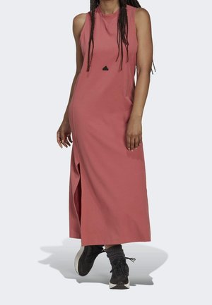 Vrouw draagt een mouwloze, enkellange roze jurk met een zijsplit en zwarte sportschoenen met grijze sokken op een lichte achtergrond.