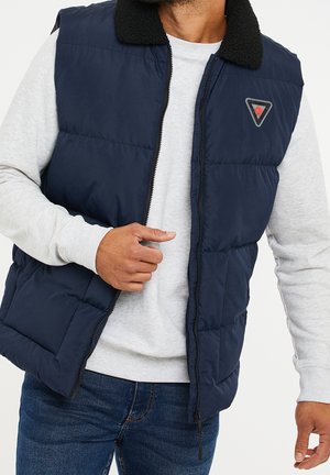 Homme portant une veste matelassée bleu marine avec col noir et logo triangulaire sur un sweat-shirt gris clair et un jean bleu, en train de fermer la veste.
