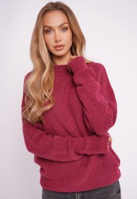 Magenta oversize’owy sweter z okrągłym dekoltem, miękką teksturą i długimi rękawami zakończonymi ściągaczami. Noszony z ciemnymi jeansami.