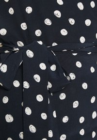 Tissu bleu marine avec des pois blancs, présentant un détail de taille nouée. La texture semble lisse et légère.