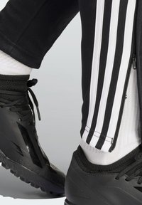 Black athletic pants with white stripes, zippered sides, paired with black sports shoes featuring textured rubber soles and adjustable laces.