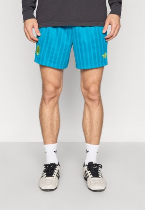 Blå shorts med lodrette striber, en afslappet pasform, med en gul logo på venstre ben. Bæres med hvide sokker og sorte sportssko.