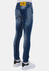 Man in slim-fit blauwe denim jeans met vervaagde delen, versleten details en witte sneakers met zwarte hakken.