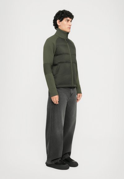 Man standing sideways wearing an olive green puffer jacket with knit sleeves, wide-leg black jeans, and black sneakers against a white background.