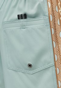 Φωτεινή γαλάζια υφασμάτινη τσέπη με μαύρη και λευκή ταινία έλξης, ραμμένες άκρες, μεταλλικό κουμπί και διπλανή καφέ διακοσμητική λωρίδα με ημικύκλια και γραμμές.