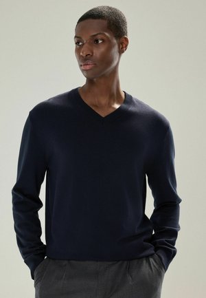 Mann trägt einen dunkelblauen V-Ausschnitt-Pullover mit den Händen in den Taschen und schaut zur Seite vor einem schlichten, hellen Hintergrund.